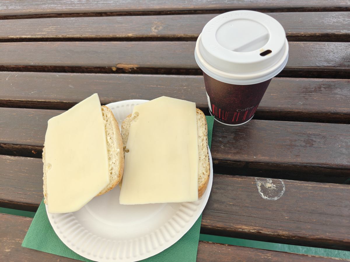 Kaffe i papkrus og et rundstykke med ost på papbakke, på en bænk ombord på færgen