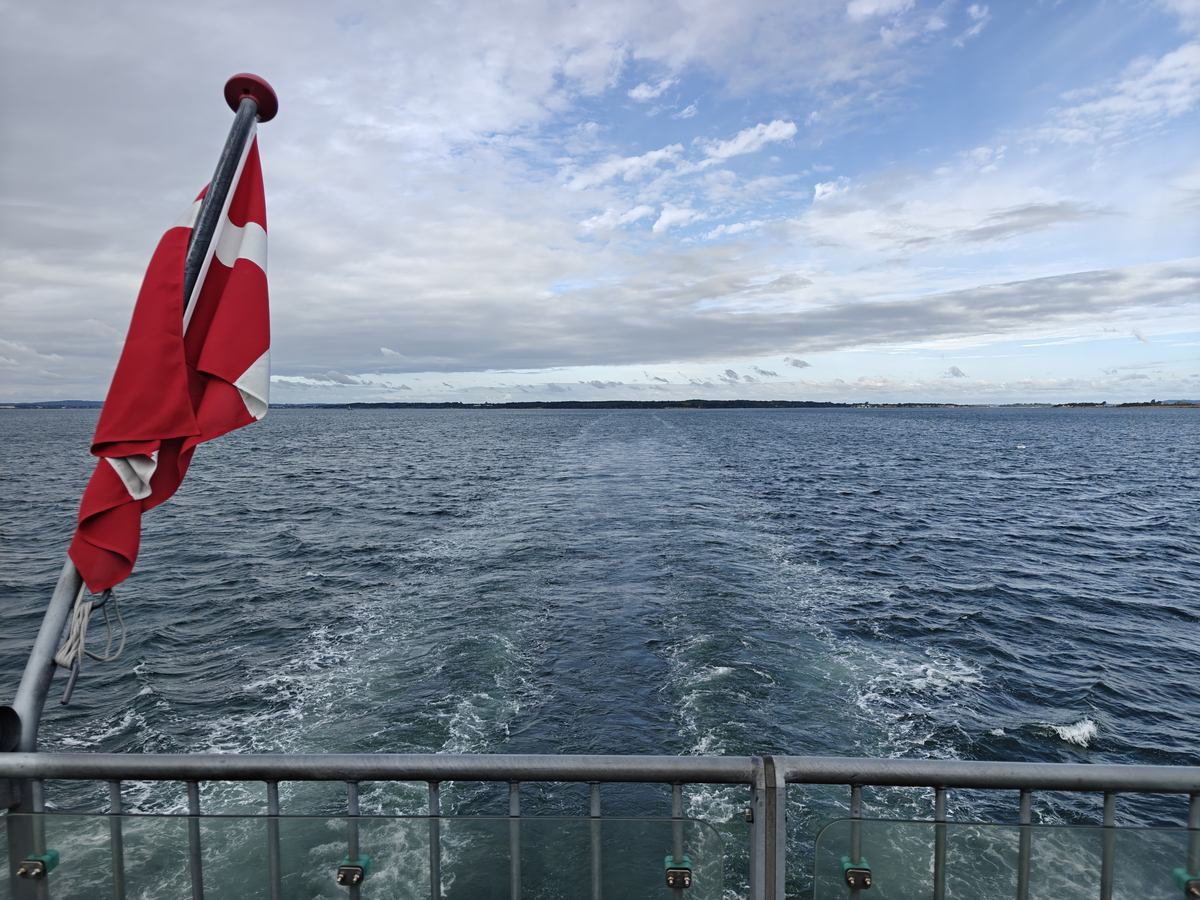 Dannebrog på en flagstang agter på færgen, med kølvandet og kysten i baggrunden