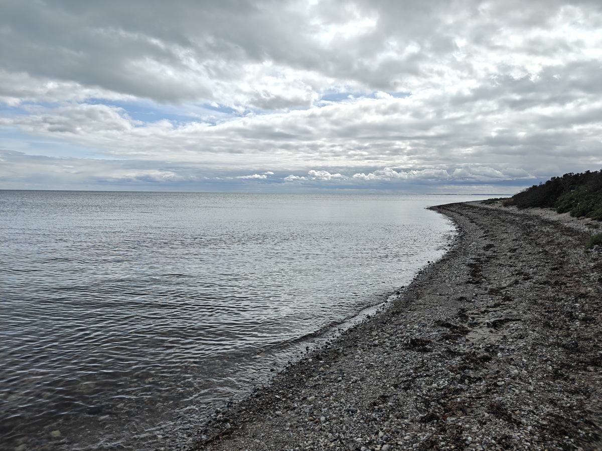 Helt stille vand ved en stenet odde, med en kystlinje i baggrunden under en overskyet himmel