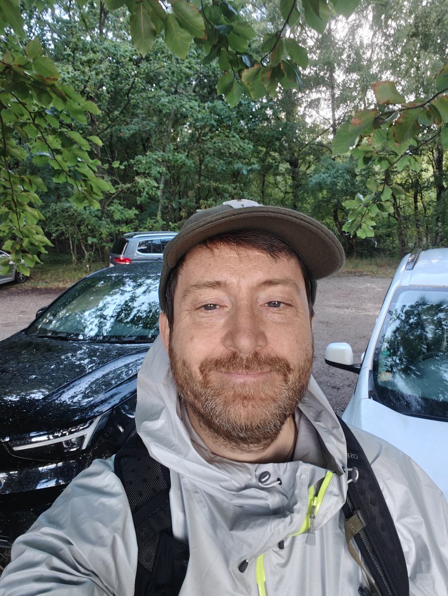Selfie med kasket og jakke foran to parkerede biler, med grønne buske i baggrunden