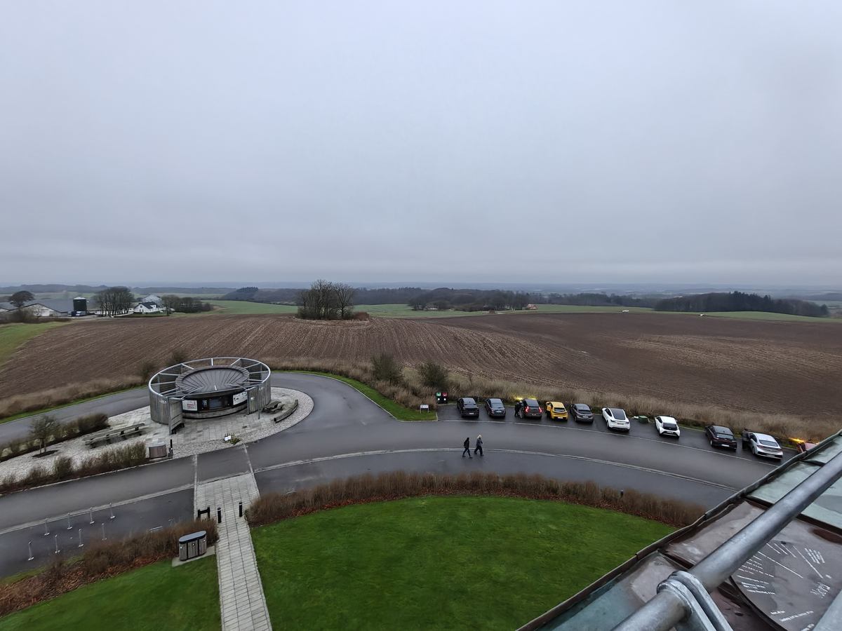 Udsigt fra toppen af tårnet ud over pløjede marker og en rundkørsel med parkerede biler, i gråvejr