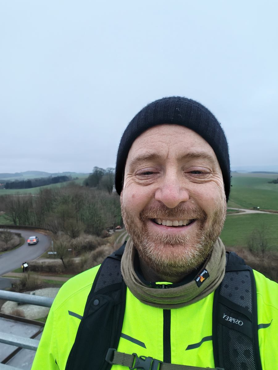 Selfie i neongul jakke og sort hue på toppen af tårnet, med marker og en vej i baggrunden